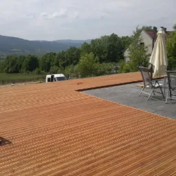 Rozległy taras drewniany z widokiem na góry, częściowo wykończony, z meblami ogrodowymi i parasolem. W tle dom i zieleń.