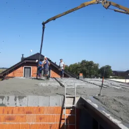 Wylewka jastrychowa na dachu domu jednorodzinnego, ekipa budowlana rozprowadza beton z pompy, widoczne bloczki ceramiczne i drabina.