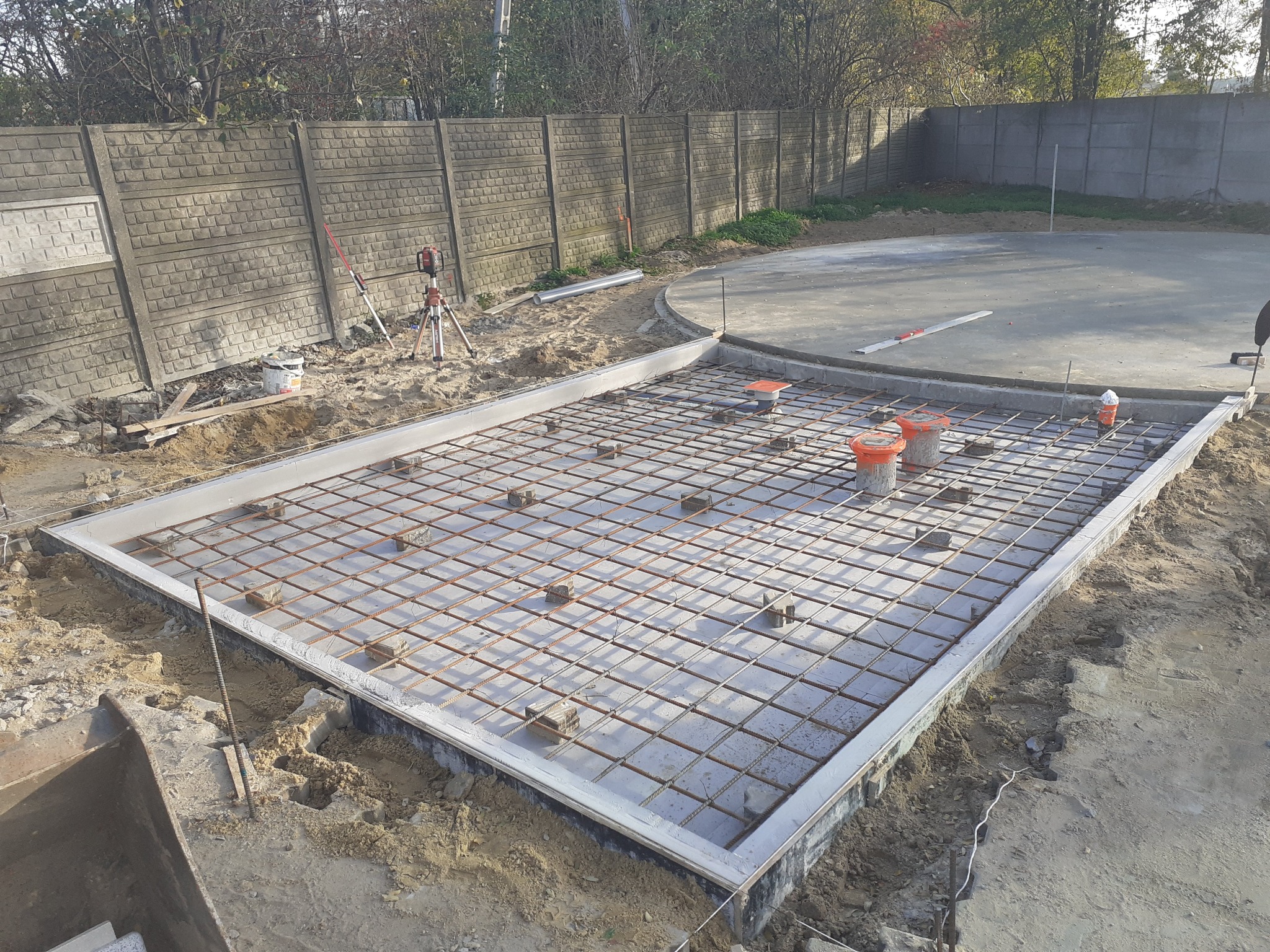 Przygotowanie zbrojenia pod wylewkę fundamentową, widoczna siatka zbrojeniowa w szalunku, obok laserowy niwelator na statywie, w tle betonowy płot.