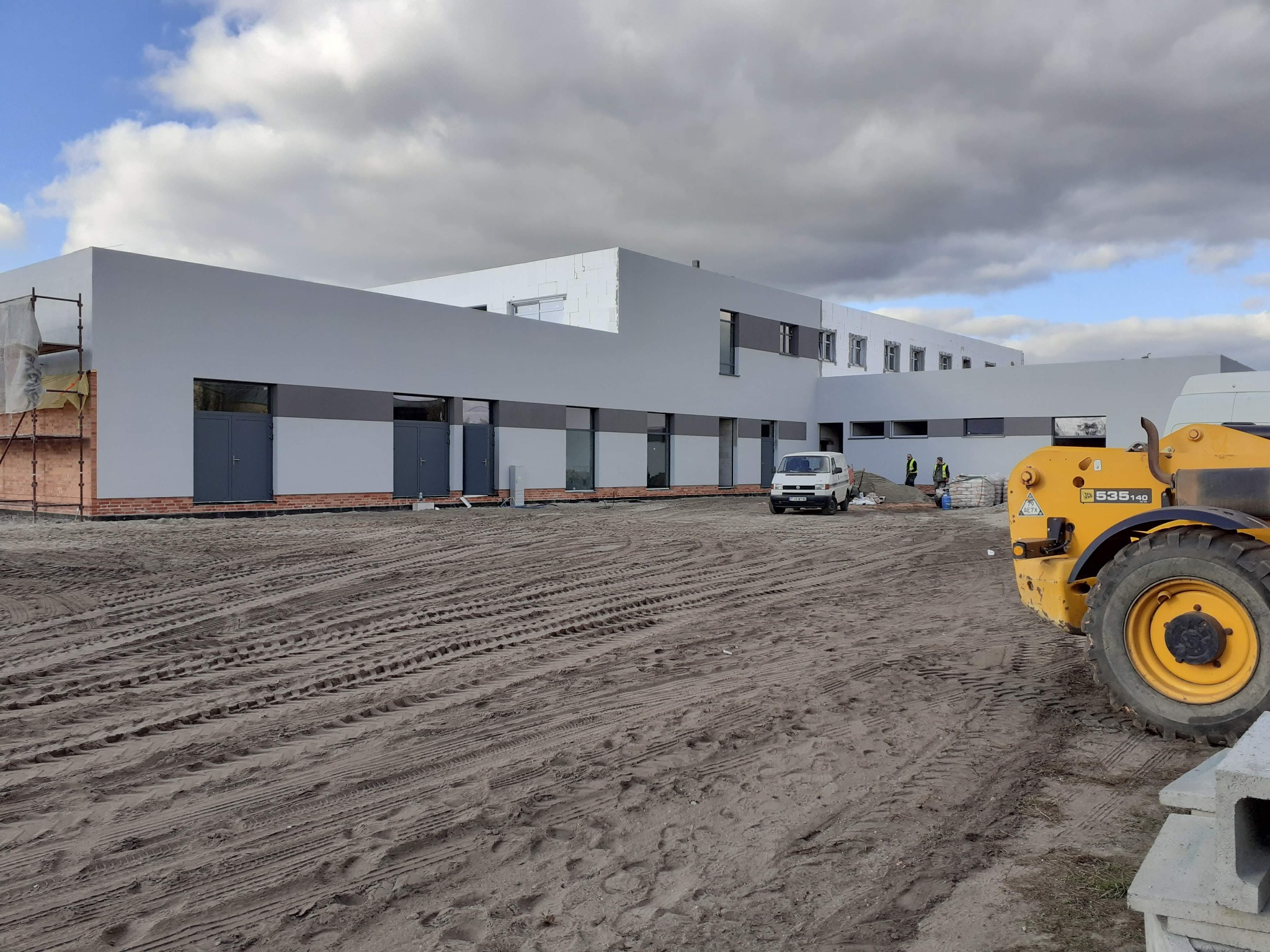 Nowoczesny budynek w trakcie wykończenia elewacji, widoczne rusztowania, niezakończone ocieplenie, teren budowy z żółtą maszyną budowlaną i śladami opon, pochmurne niebo.