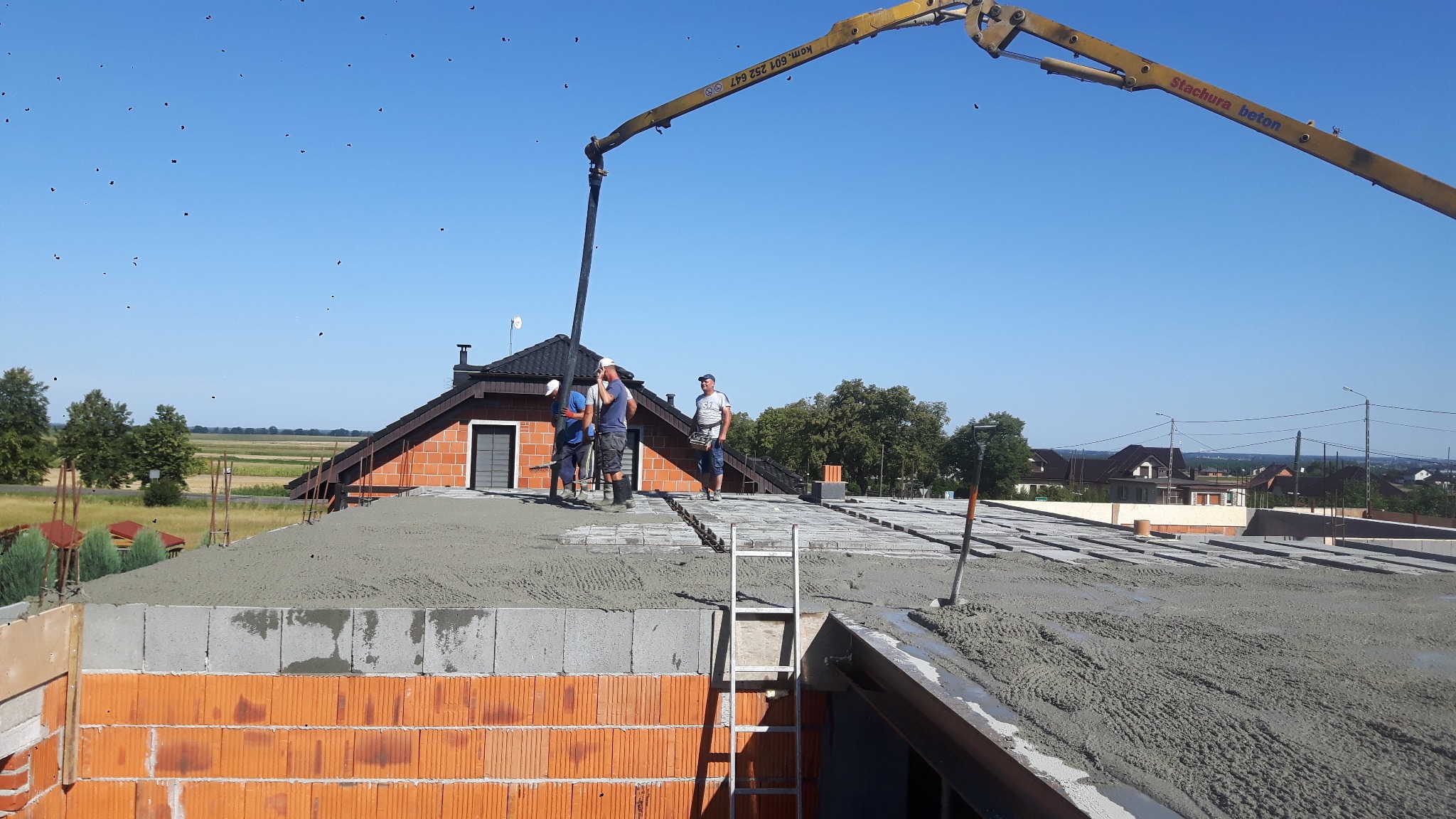 Wylewka jastrychowa na dachu domu jednorodzinnego, ekipa budowlana rozprowadza beton z pompy, widoczne bloczki ceramiczne i drabina.