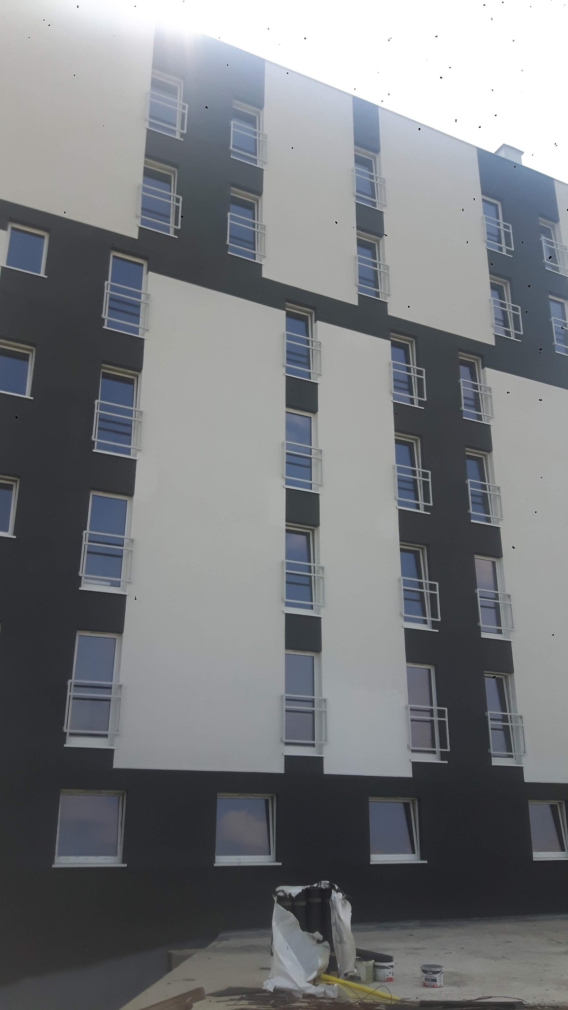 Wielopiętrowy budynek z elewacją w kolorach biało-grafitowych, widoczne puszki z farbą i folia malarska na dole, elewacja z balkonami.