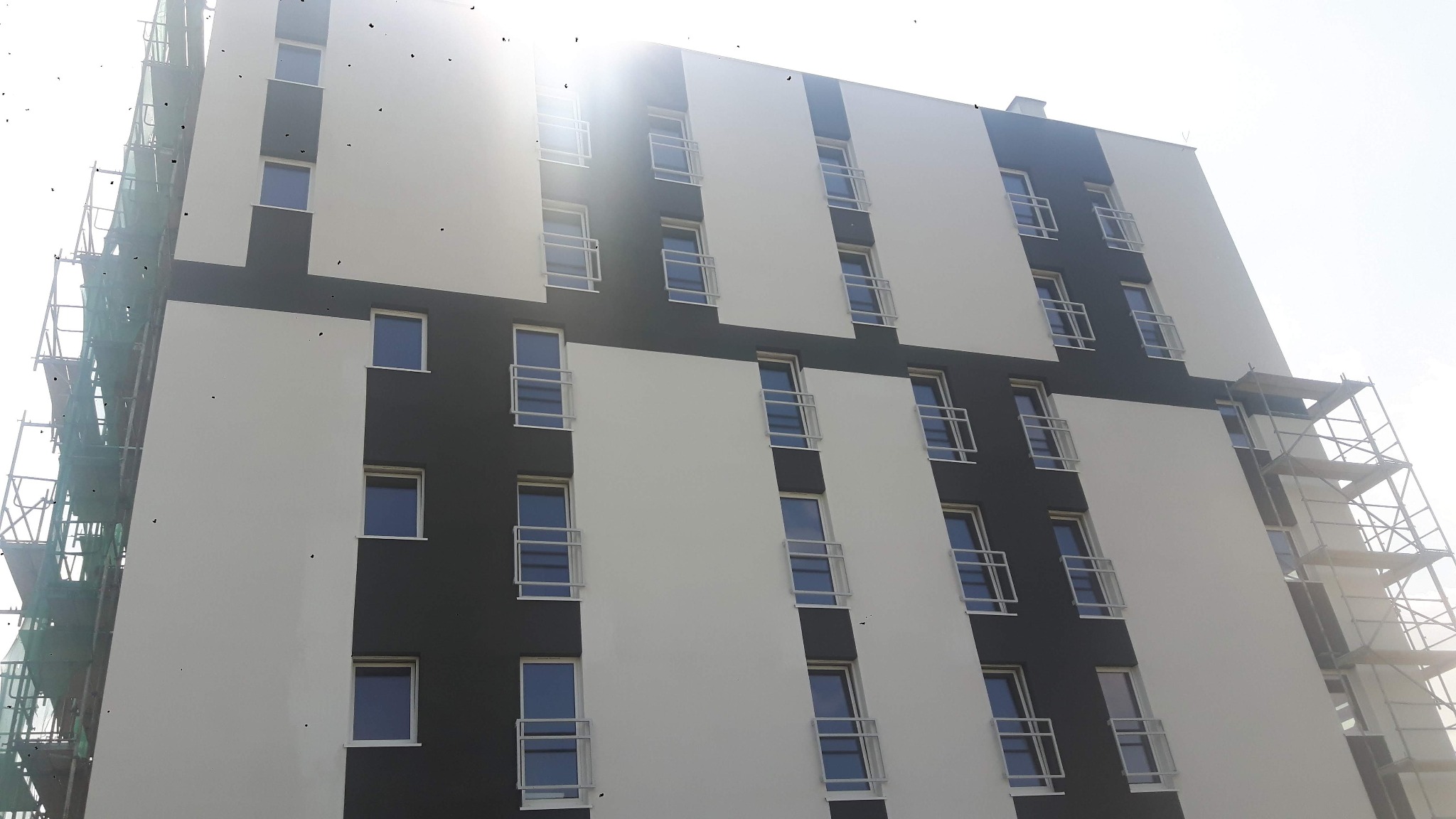 Nowoczesny budynek wielopiętrowy z biało-szarą elewacją, widoczne rusztowania po lewej stronie i w prawym górnym rogu, oświetlony silnym słońcem.
