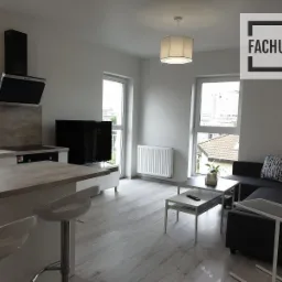 Jasne, nowocześnie urządzone mieszkanie z aneksem kuchennym, wyspą z hokerami, szarą sofą z wzorzystymi poduszkami i widokiem z okna na zabudowę miejską, z logo firmy w rogu.