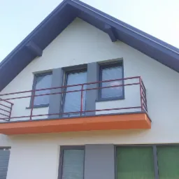 Elewacja domu jednorodzinnego z balkonem, wykończona w jasnych kolorach z grafitowymi detalami dachu i okien. Balkon z pomarańczową podłogą i bordową barierką.