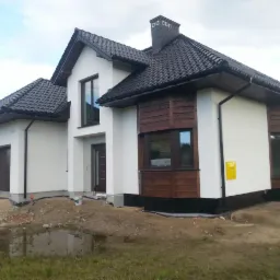 Nowoczesny dom jednorodzinny w Radgoszczu z białą elewacją, drewnianymi akcentami i ciemną dachówką. Widoczny fragment terenu przed budynkiem.