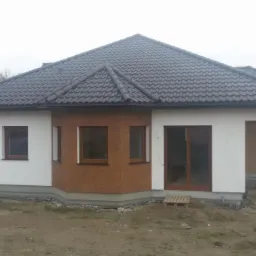 Dom jednorodzinny z dachem z ciemnej dachówki, częściowo otynkowany na biało, a częściowo wykończony panelami w kolorze drewna. Widoczny pracownik w oddali.