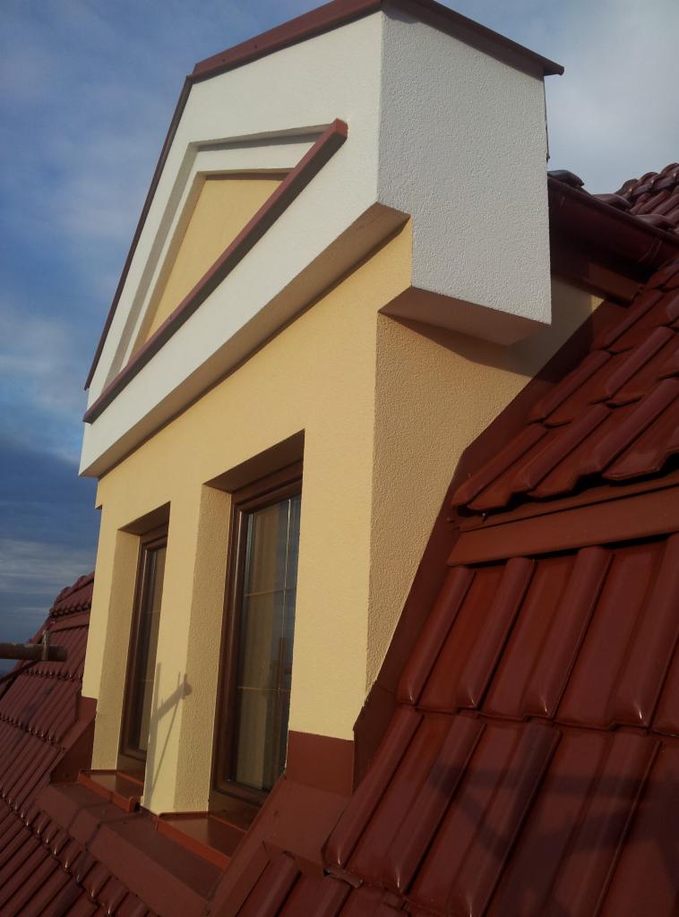 Wykończona lukarna z oknami na dachu pokrytym czerwoną dachówką. Elewacja w odcieniach beżu i bieli. Widok z dołu, podkreślający detale architektoniczne.