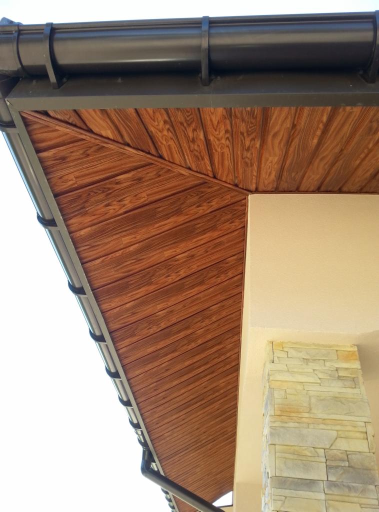 Podbitka dachowa z drewnianym wzorem, brązowe rynny i fragment elewacji z kamienną kolumną. Ujęcie z dołu, widoczne detale wykończenia.