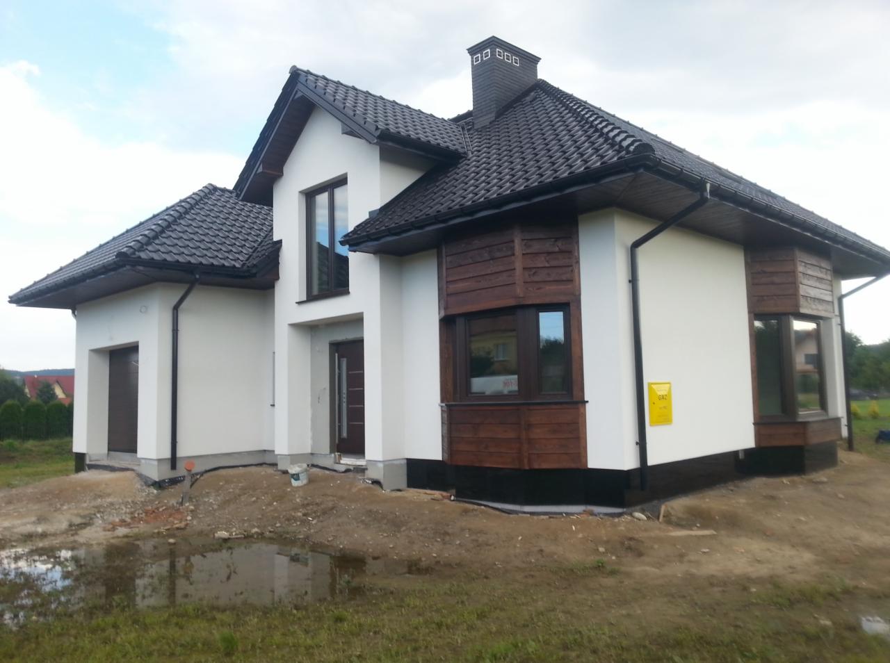 Nowoczesny dom jednorodzinny w Radgoszczu z białą elewacją, drewnianymi akcentami i ciemną dachówką. Widoczny fragment terenu przed budynkiem.