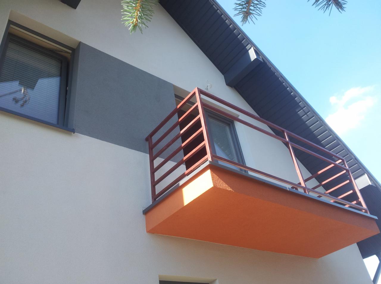 Elewacja domu z balkonem po remoncie: biała ściana, szary pas obok okna, pomarańczowy balkon z brązową balustradą. Fragment dachu i iglastej gałązki w tle.