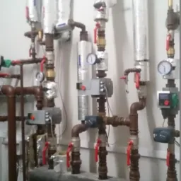 Rozbudowana instalacja z rur miedzianych i zaworów z czerwonymi uchwytami, manometry i sterowniki przy białej ścianie, izolowane termicznie pionowe rury