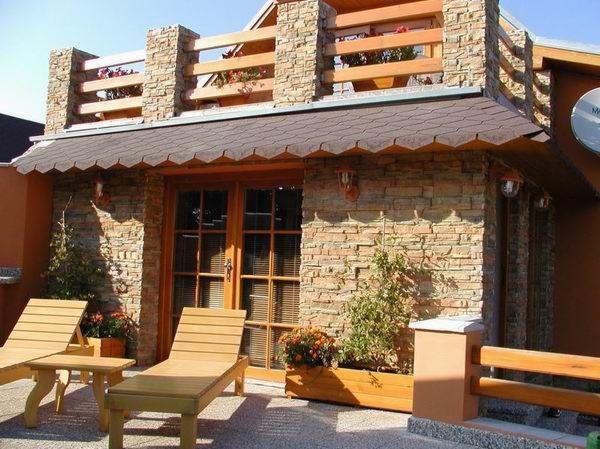 Elewacja budynku z kamienia dekoracyjnego, drewniane okna i balkon z kwiatami. Dwa drewniane leżaki na tarasie. Ciepłe oświetlenie podkreśla fakturę kamienia.