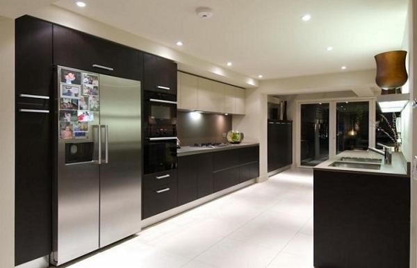 Nowoczesna, przestronna kuchnia z ciemnymi szafkami, stalową lodówką z magnesami i białymi płytkami podłogowymi. Widoczne duże okno i oświetlenie punktowe.