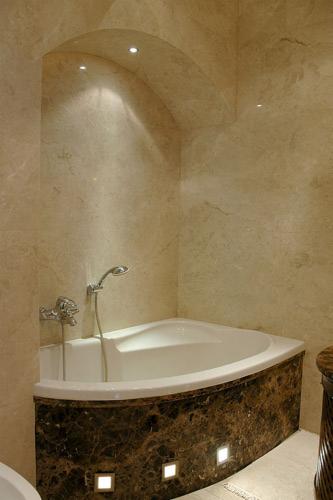 Elegancka narożna wanna z marmurową obudową i podświetleniem LED w łazience wykończonej jasnym trawertynem, oświetlona punktowymi lampami sufitowymi.