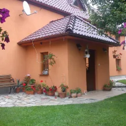 Pomarańczowy dom z brązowym dachem, widok z ogrodu, z kwiatami w doniczkach na kamiennym podłożu i drewnianą ławką z boku.