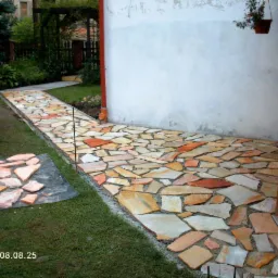 Ścieżka ogrodowa wyłożona nieregularnymi płytami z piaskowca w odcieniach beżu i czerwieni, prowadząca wzdłuż białej ściany budynku, z fragmentem przygotowanych płyt na trawie.