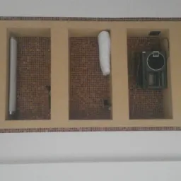 Nietypowe wnęki w łazience wykończone mozaiką, z dekoracyjnymi elementami. Widoczne rury i sprzęt elektroniczny. Całość w ciepłej tonacji beżu i brązu.