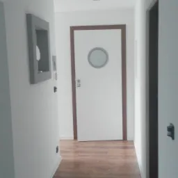 Białe drzwi z okrągłym oknem w drewnianej ramie, widoczne z perspektywy korytarza z drewnianą podłogą i szarą dekoracją ścienną.