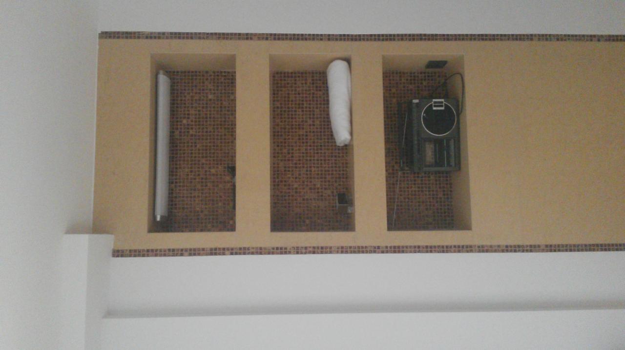 Nietypowe wnęki w łazience wykończone mozaiką, z dekoracyjnymi elementami. Widoczne rury i sprzęt elektroniczny. Całość w ciepłej tonacji beżu i brązu.