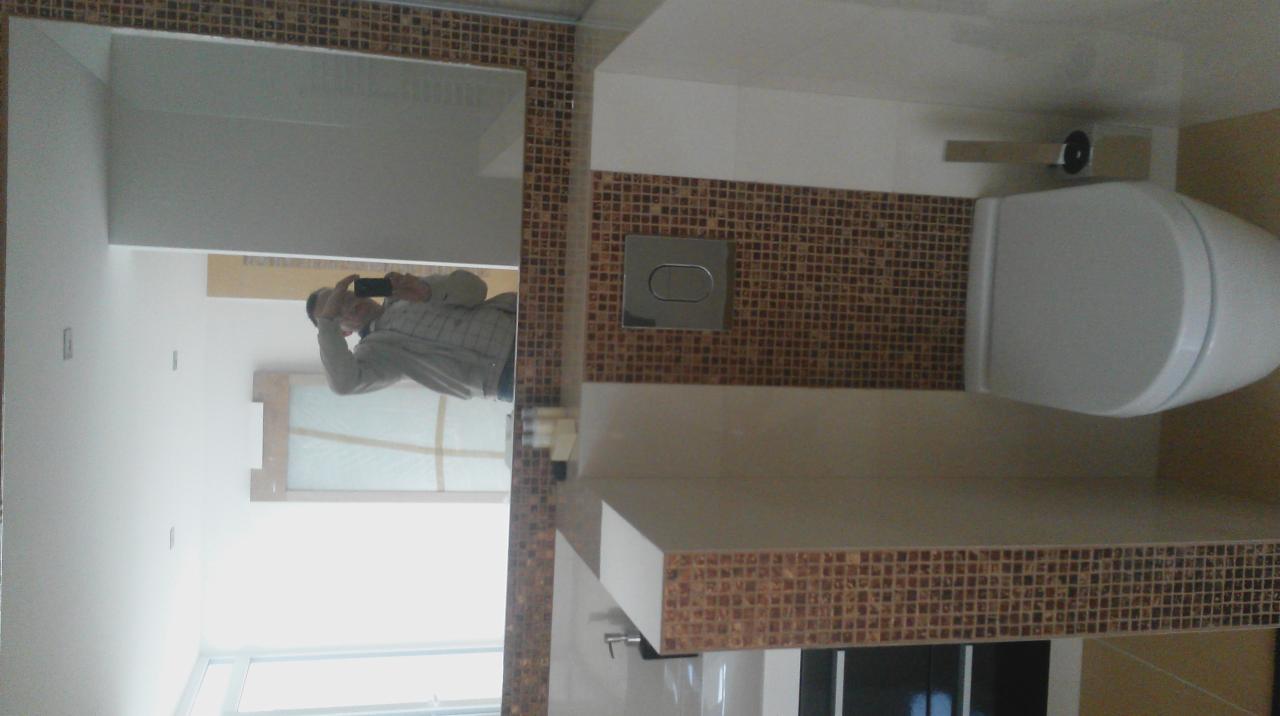 Wykończona toaleta z mozaiką w odcieniach brązu, widok z góry. W lustrze odbija się osoba robiąca zdjęcie. Minimalistyczny design.