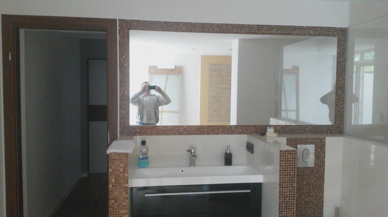 Wykończona łazienka z umywalką i lustrem w mozaikowej ramie. W lustrze odbija się osoba robiąca zdjęcie telefonem. Widoczne elementy armatury i kosmetyki.