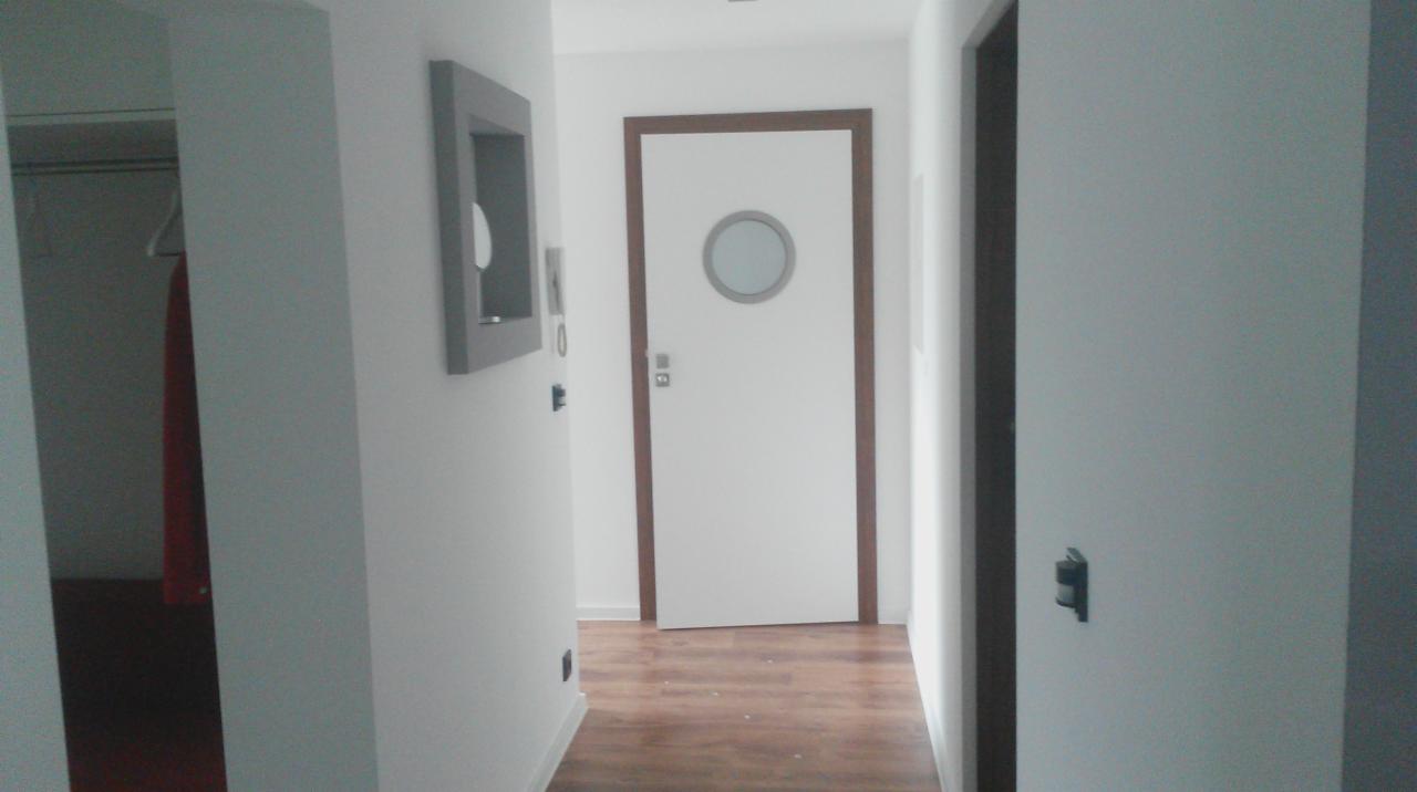 Białe drzwi z okrągłym oknem w drewnianej ramie, widoczne z perspektywy korytarza z drewnianą podłogą i szarą dekoracją ścienną.