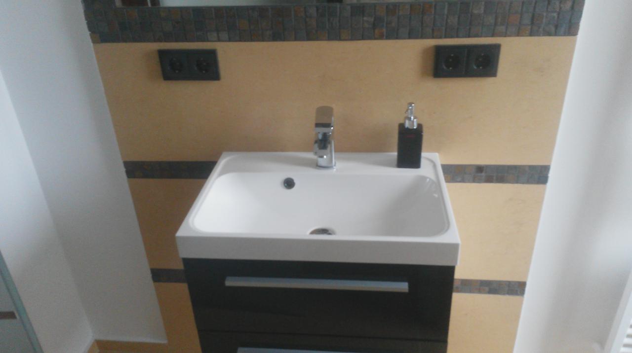 Umywalka z szafką w nowoczesnej łazience, beżowe ściany z mozaiką, chromowana bateria i czarny dozownik mydła. Widoczne gniazdka elektryczne.