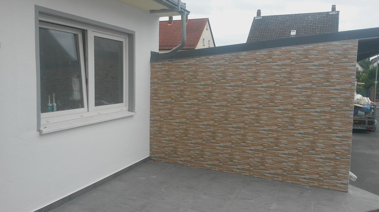 Wykończony balkon z szarą podłogą i dekoracyjną ścianą z płytek imitujących kamień. Widoczne okno i fragment dachu sąsiedniego budynku.