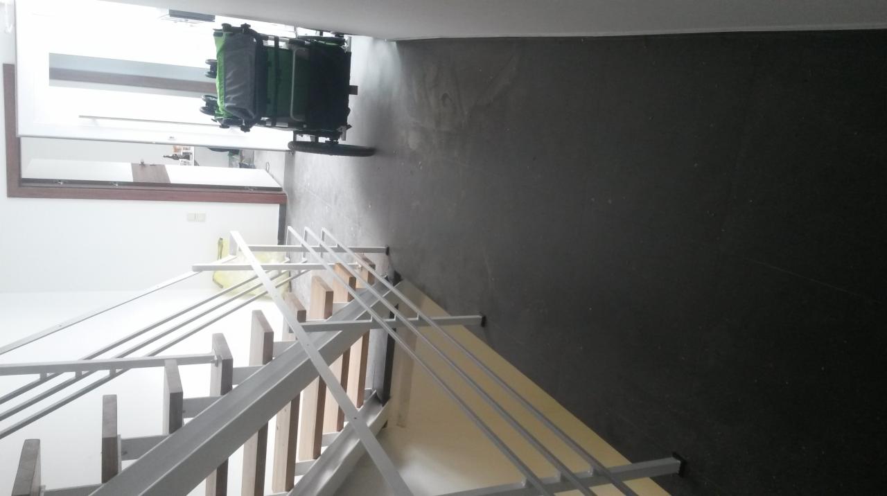 Białe, metalowe schody z drewnianymi stopniami w jasnym wnętrzu. Widoczny fragment podłogi i ściany, a także wózek transportowy w tle.