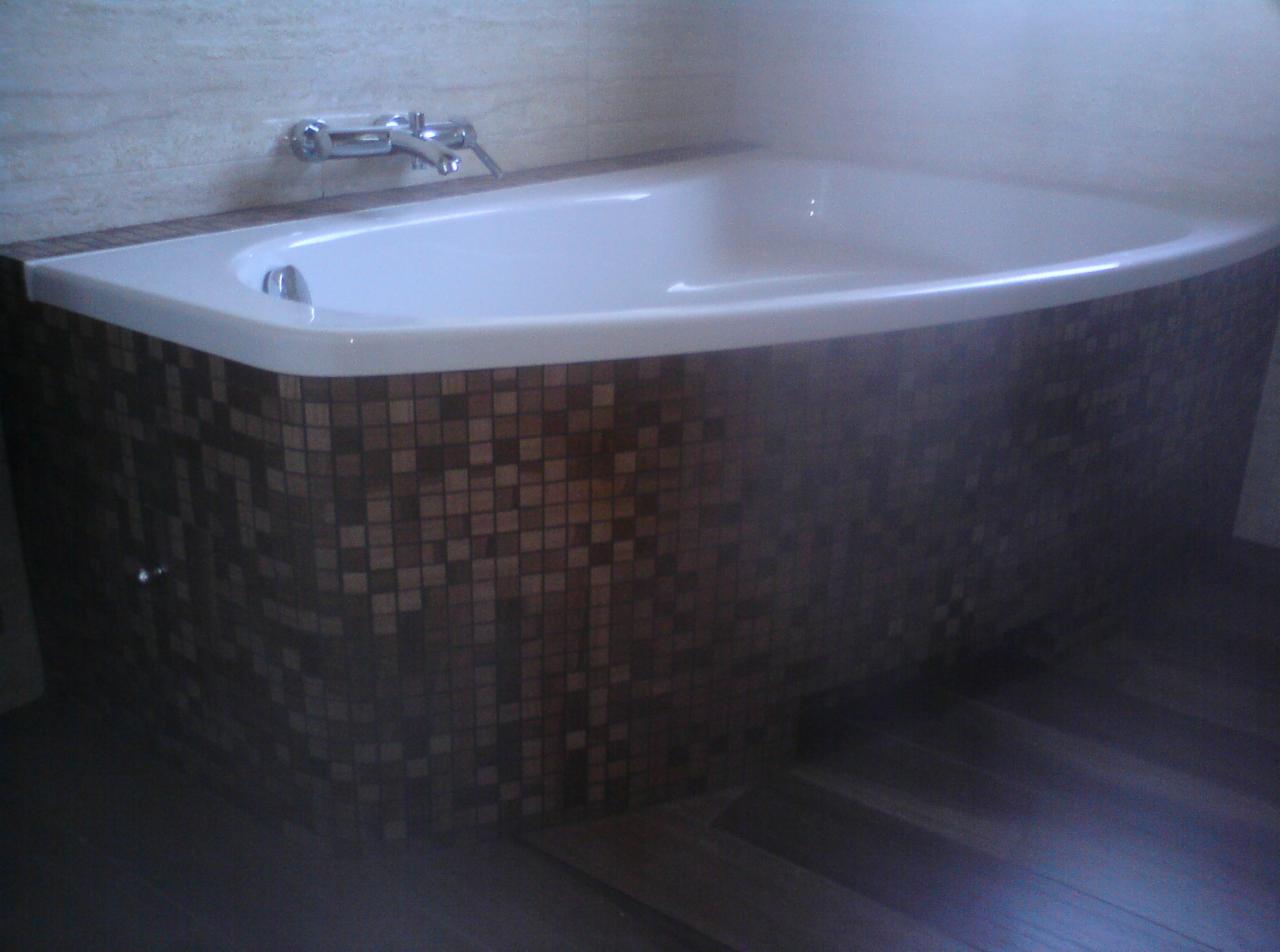 Biała wanna narożna obudowana mozaiką w odcieniach brązu, z chromowaną baterią. Na podłodze ciemne panele. Ściana wyłożona jasnymi płytkami.