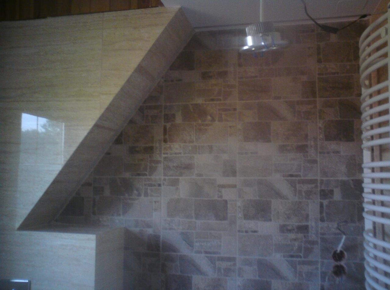 Ukończona ściana łazienki z beżowych płytek o nieregularnym kształcie, oświetlenie sufitowe i biały grzejnik drabinkowy po prawej stronie.