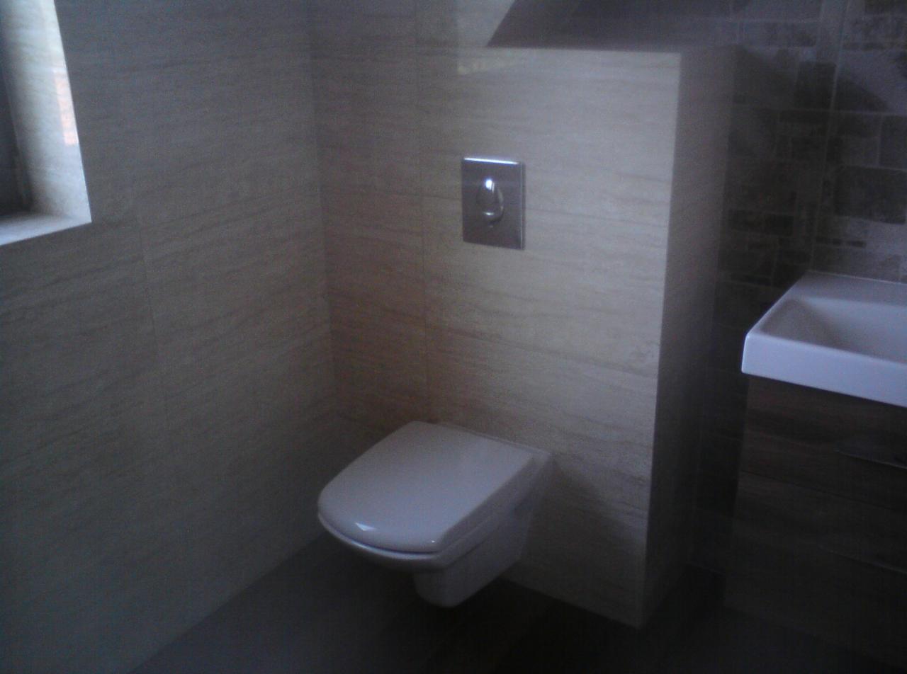 Nowoczesna łazienka z podwieszaną toaletą i umywalką w odcieniach beżu i brązu. Minimalistyczny design, płytki imitujące drewno, chromowany przycisk spłukiwania.