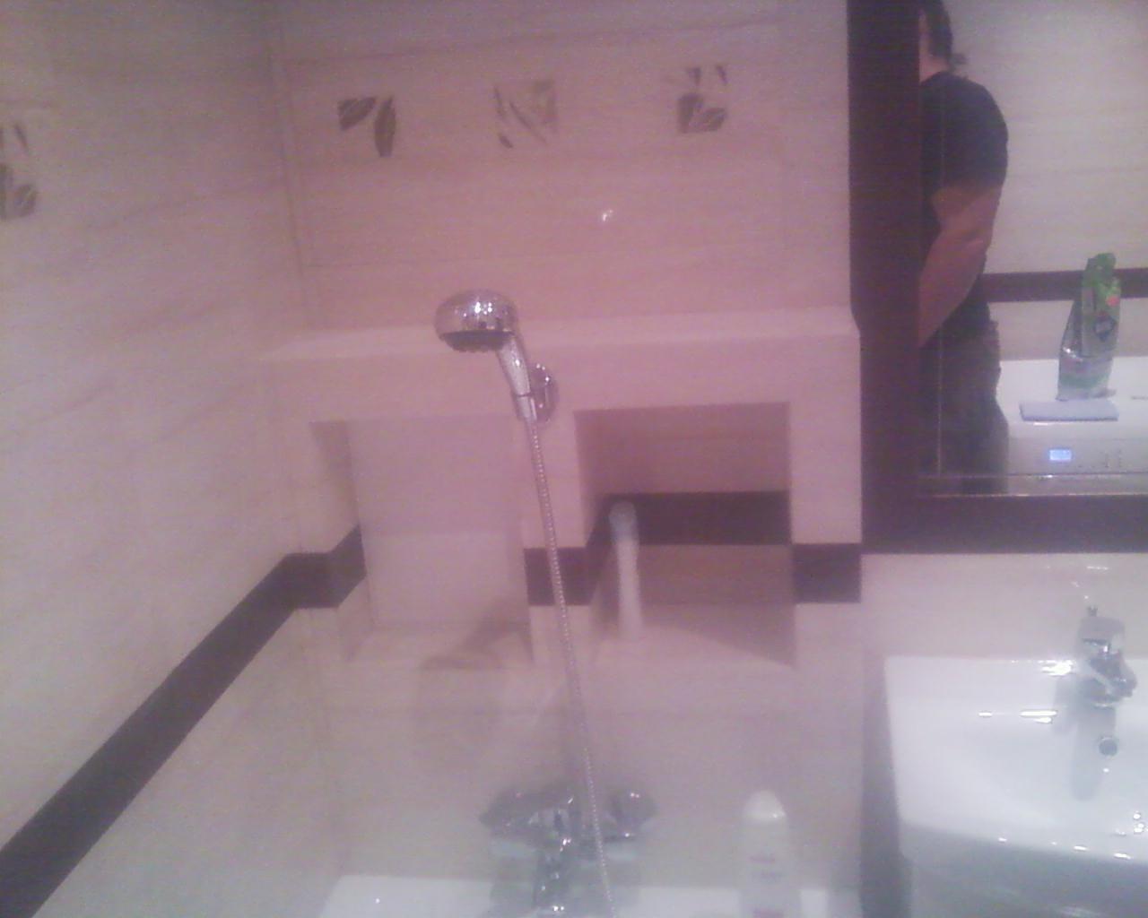 Wykończona łazienka z wanną, prysznicem i umywalką. Na ścianach jasne płytki z delikatnym motywem, ciemny pas płytek. Widoczne odbicie osoby w lustrze.