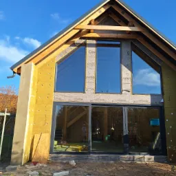 Dom w trakcie ocieplania, widoczne płyty izolacyjne na elewacji, okna z refleksami odbijającymi wnętrze, rusztowanie obok ściany, nieotynkowany fragment elewacji.