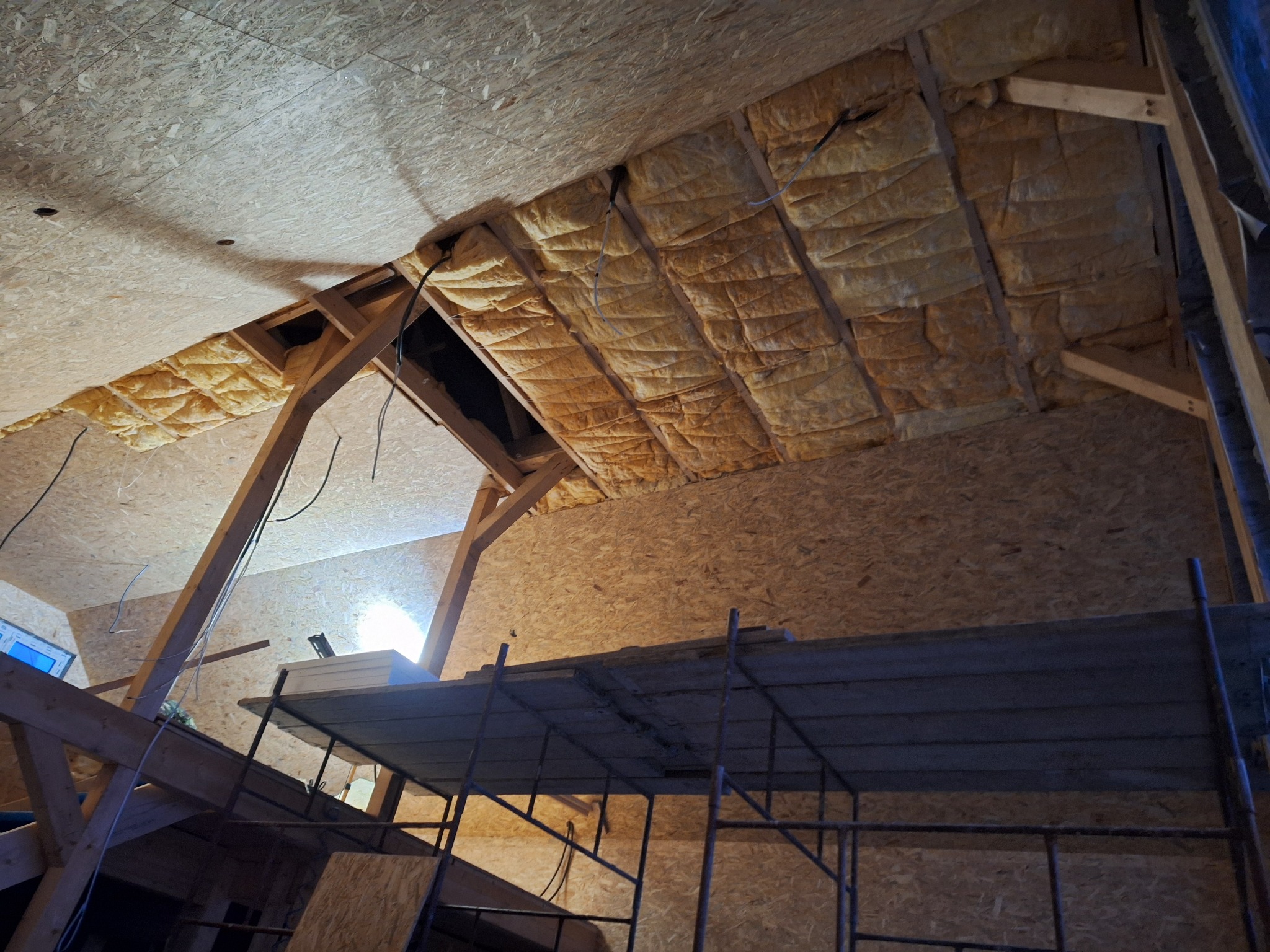 Wnętrze poddasza w trakcie prac: widoczne ocieplenie wełną mineralną między krokwiami, płyty OSB na ścianach, drewniana konstrukcja oraz metalowe rusztowanie.