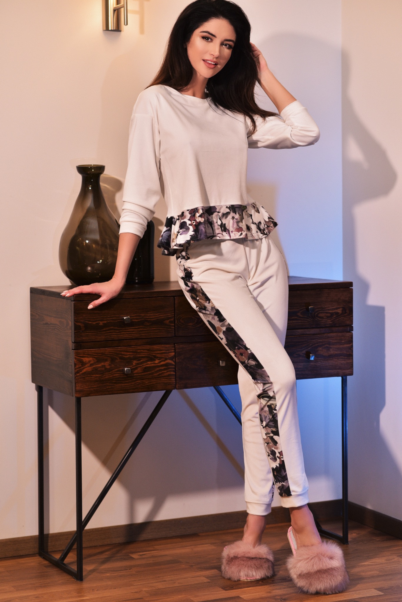 Kobieta w białym dresie z kwiatowym wzorem i futrzanych kapciach opiera się o drewnianą komodę z ciemnego drewna, obok wazon.