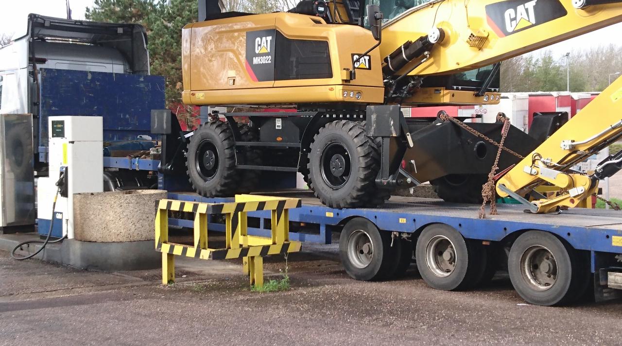 Żółta koparka CAT MH3022 na niebieskiej lawecie, obok dystrybutor paliwa i betonowy blok, w tle drzewa.