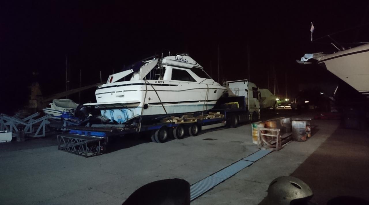 Biała motorówka 'Lambada' z numerem rejestracyjnym DL 4814 W, zabezpieczona na niskopodwoziowej platformie transportowej z ciężarówką w tle, nocne ujęcie w porcie.