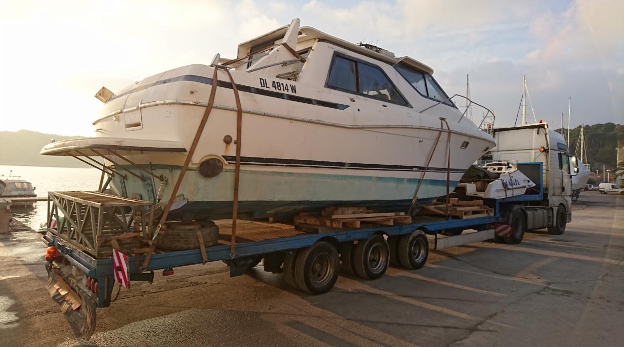 Transport jachtu na niskopodwoziowej naczepie. Widoczny numer rejestracyjny jachtu. W tle marina i woda. Słoneczny dzień.