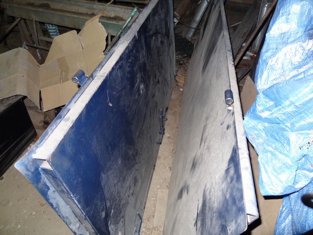 Dwie duże, metalowe płyty, prawdopodobnie elementy bramy, ustawione pionowo obok siebie w zagraconym pomieszczeniu. Widoczne zawiasy i metalowe ramy.