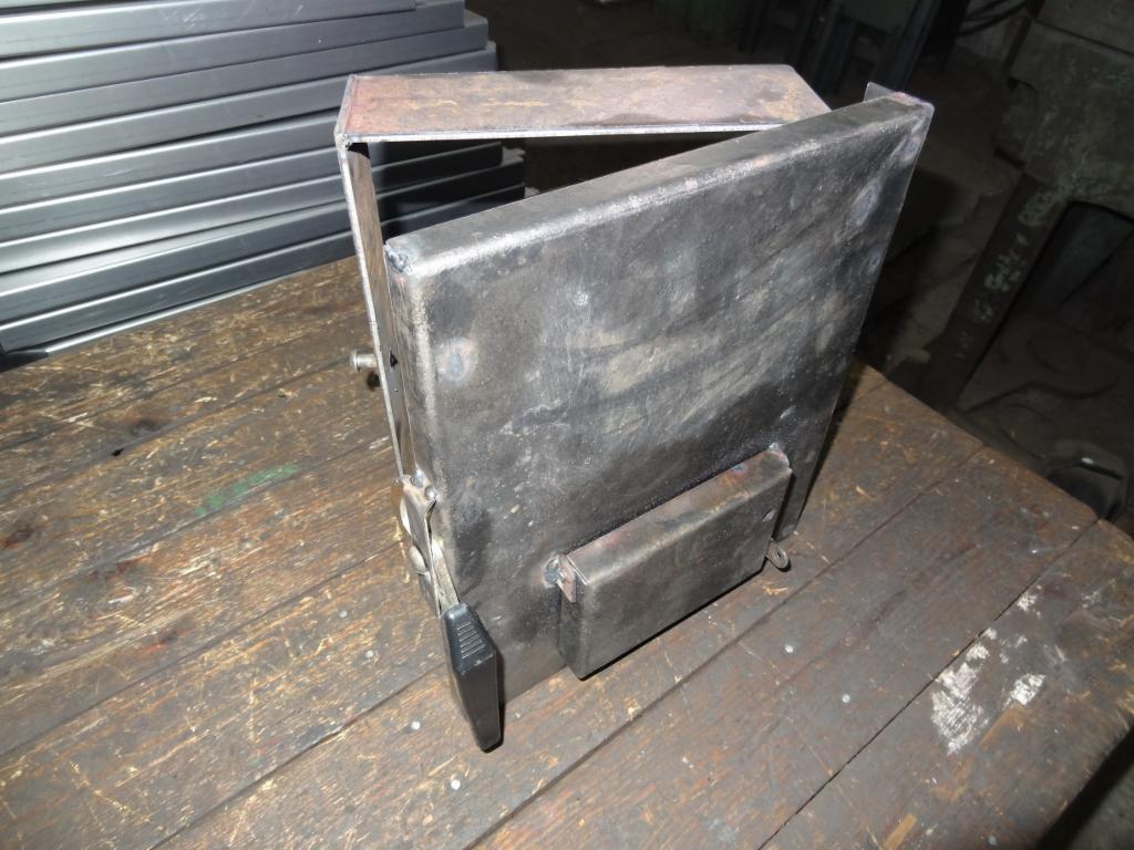 Metalowa konstrukcja z otwieranymi drzwiczkami, spawana, umieszczona na drewnianym blacie. W tle widoczne metalowe profile.