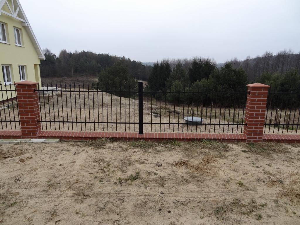 Czarne, metalowe ogrodzenie z ceglanymi słupkami i podmurówką na piaszczystym terenie, z widokiem na las w tle i fragmentem żółtego domu po lewej stronie.
