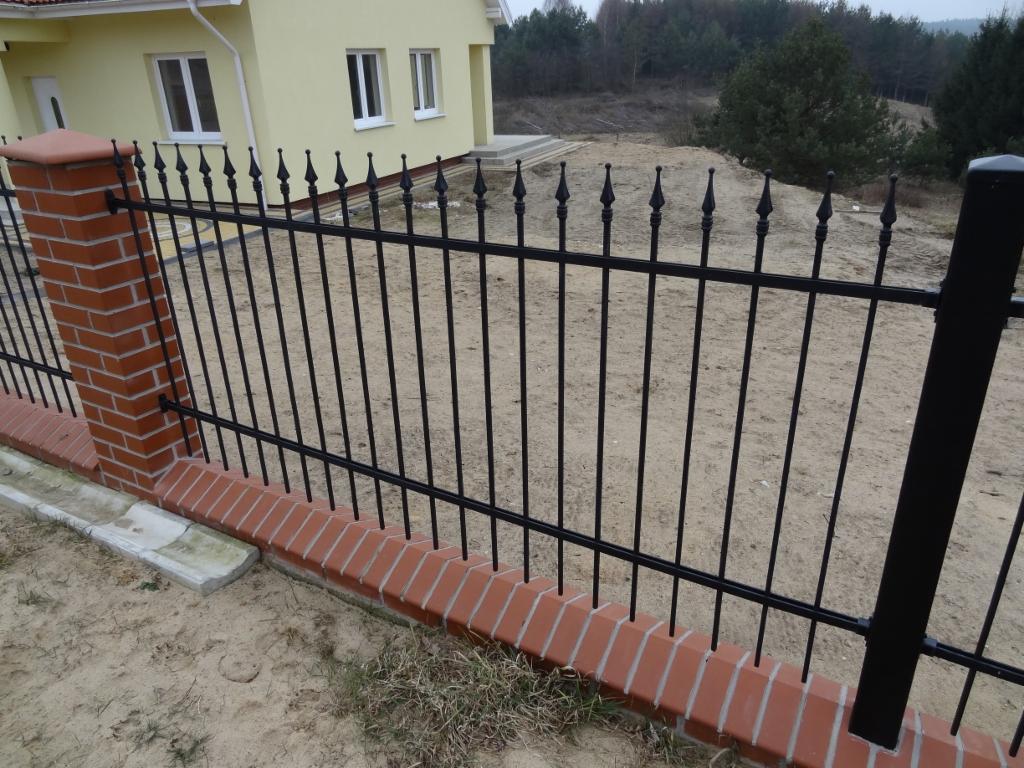 Czarne, metalowe ogrodzenie z ceglanymi słupkami na tle żółtego domu i piaszczystego terenu. Szpiczaste zakończenia prętów dodają ogrodzeniu dekoracyjnego charakteru.