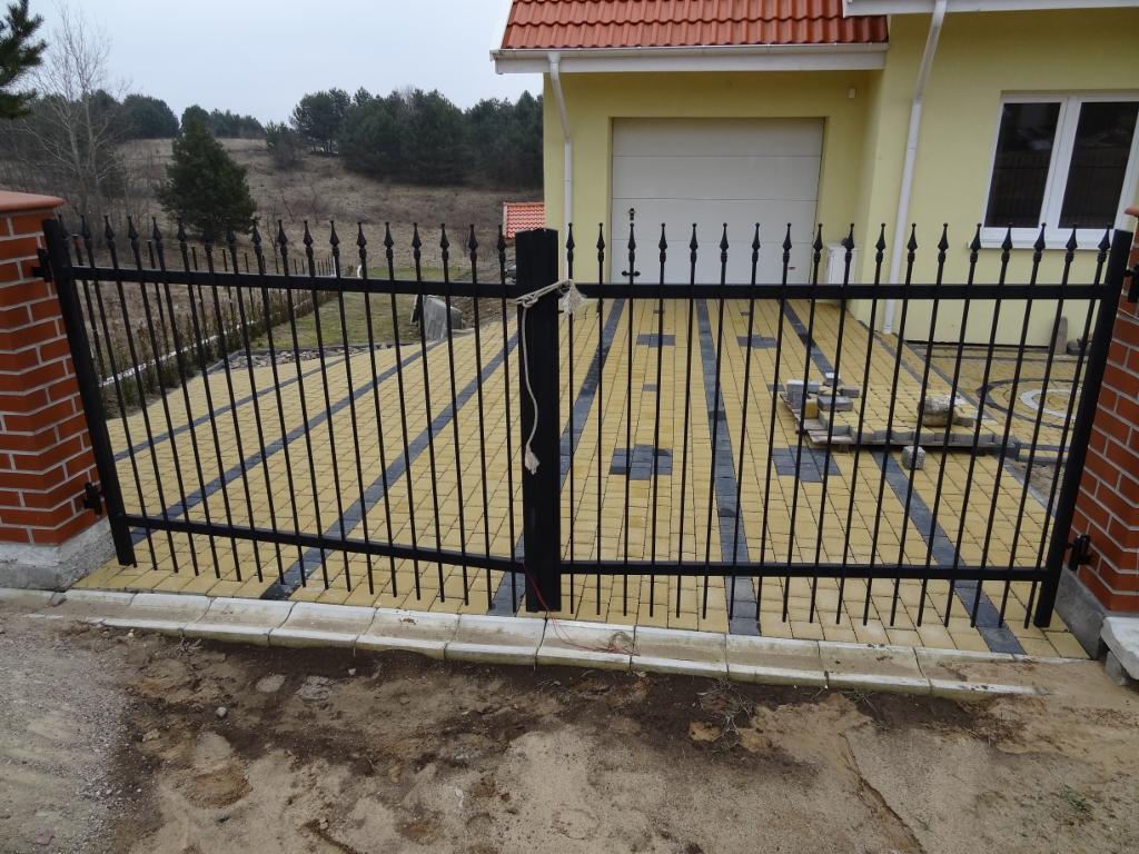 Czarna, metalowa brama wjazdowa z dekoracyjnymi grotami, zamontowana na ceglanych słupkach. W tle widoczny dom z garażem i podjazd z kostki.
