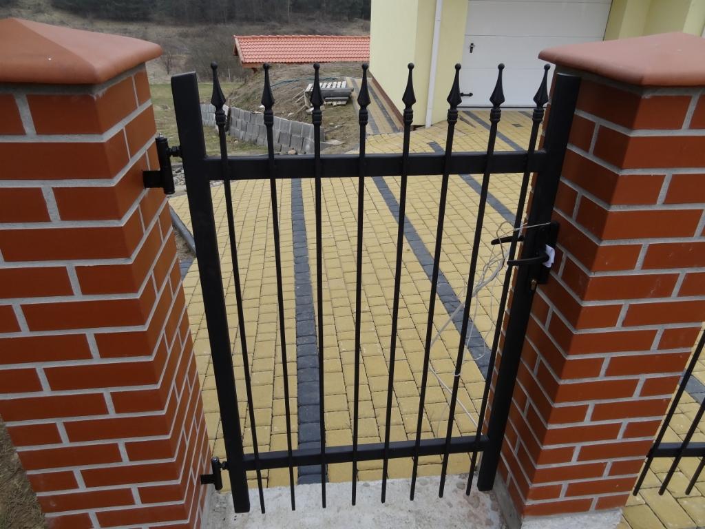 Kuta, czarna furtka z ozdobnymi grotami, osadzona między dwoma ceglanymi słupkami. W tle kostka brukowa i budynek z białą bramą garażową.