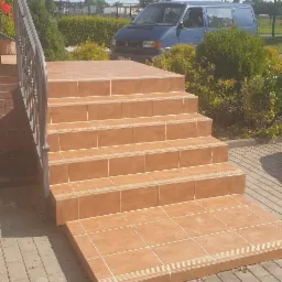 Wykończone schody wejściowe wyłożone terakotą z metalową barierką, na zewnątrz kostka brukowa, w tle zaparkowany niebieski van.