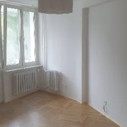 Pusty pokój po remoncie z parkietem w jodełkę, białymi ścianami i grzejnikami pod oknem, widok na zwisającą lampę z abażurem.