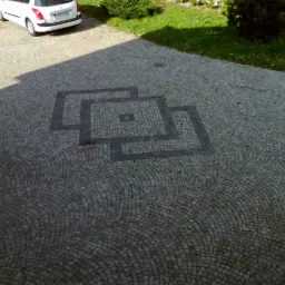 Granitowa kostka brukowa ułożona w łukowaty wzór z ciemniejszym geometrycznym motywem. W tle zaparkowany biały samochód i fragment zielonego trawnika.