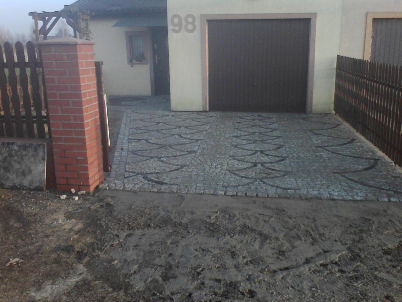 Ułożona kostka granitowa w kształcie wzoru przed garażem i domem z numerem 98, widoczny fragment ogrodzenia z ceglanym słupkiem.